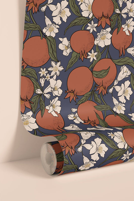 A roll of wallpaper with a repeating pattern of pomegranates, white flowers, and green leaves on a dark blue background, shown partially unrolled against a warm beige wall.
