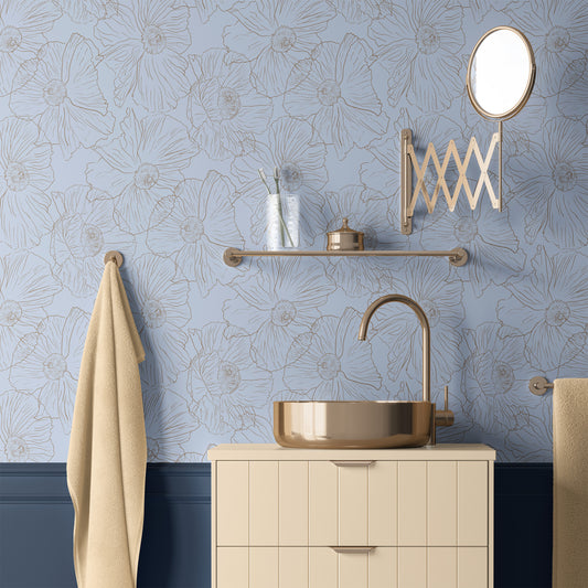 A styled bathroom scene featuring light blue floral wallpaper with a beige vessel sink on a cream vanity, a gold faucet, a wall-mounted gold mirror, and a beige towel hanging on a brass ring.