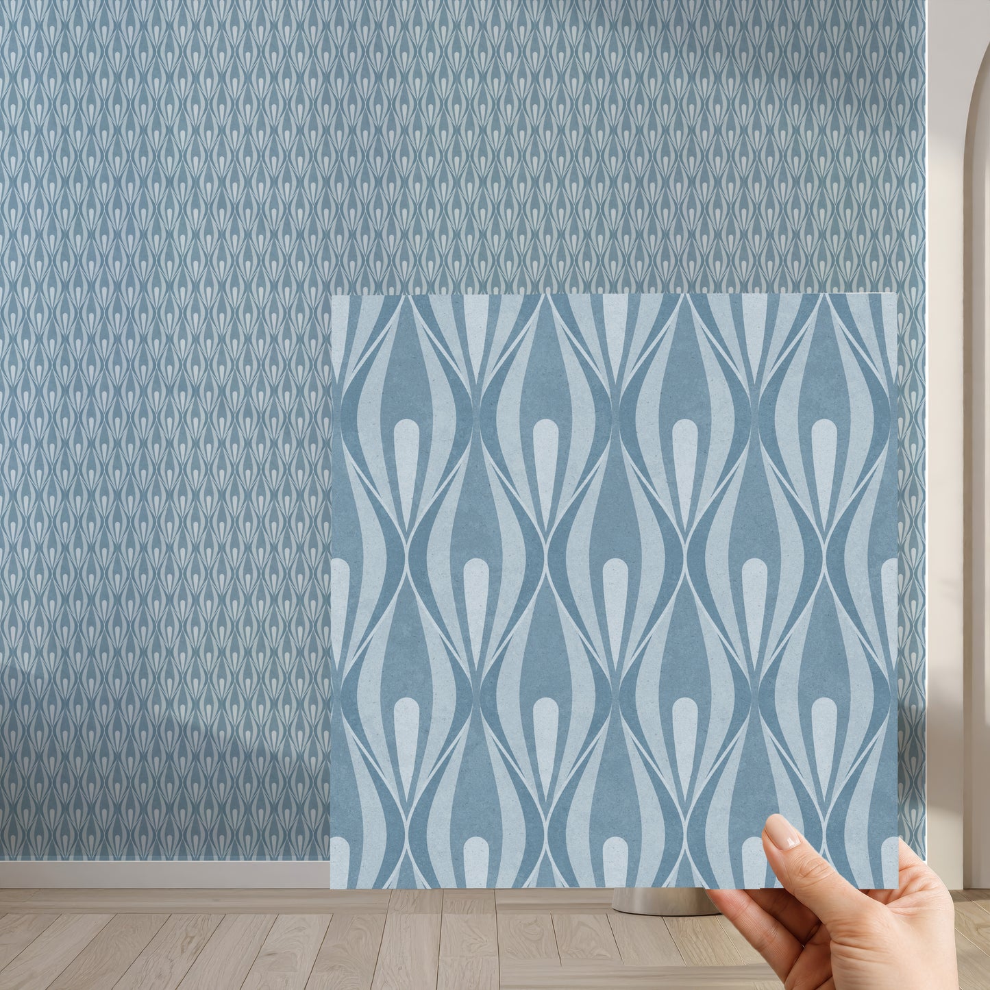 A hand holds a square sample of wallpaper with a light blue wavy geometric pattern in front of a larger wall showing a denser blue 70s ogee pattern.