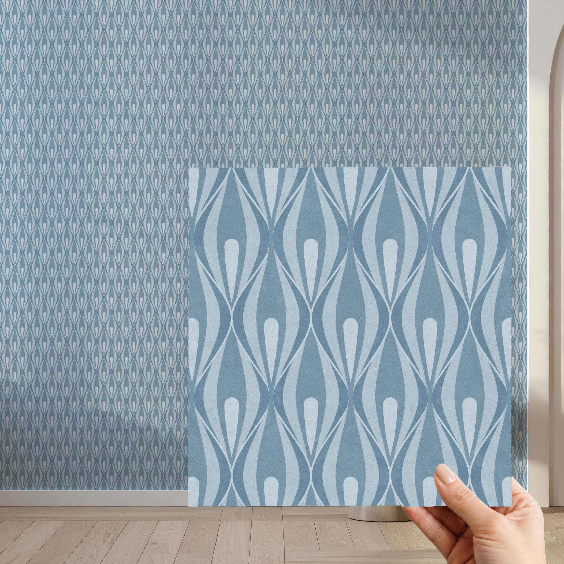 A hand holds a square sample of wallpaper with a light blue wavy geometric pattern in front of a larger wall showing a denser blue 70s ogee pattern.