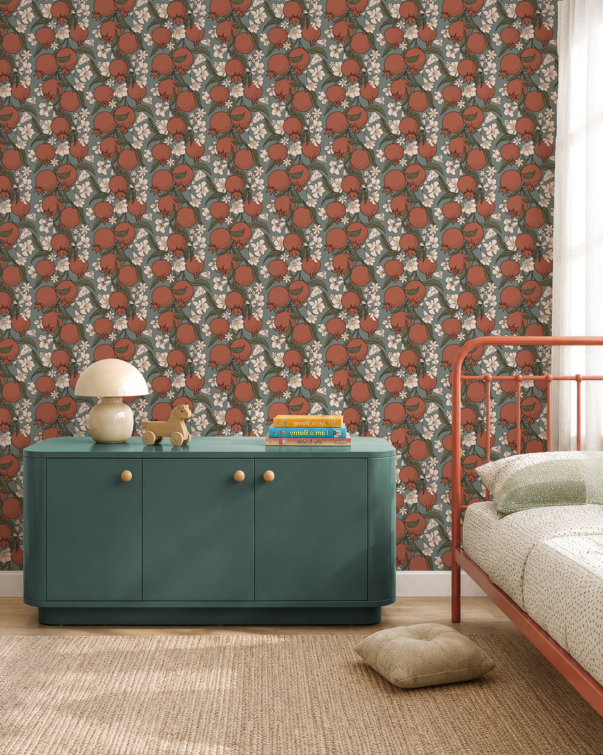 A styled bedroom corner featuring a dark green wallpaper with a repeating pattern of red-orange pumpkins and white flowers. A teal green sideboard cabinet with gold knobs sits against the wall, styled with a mushroom lamp and books, next to a rust-orange metal bed frame.