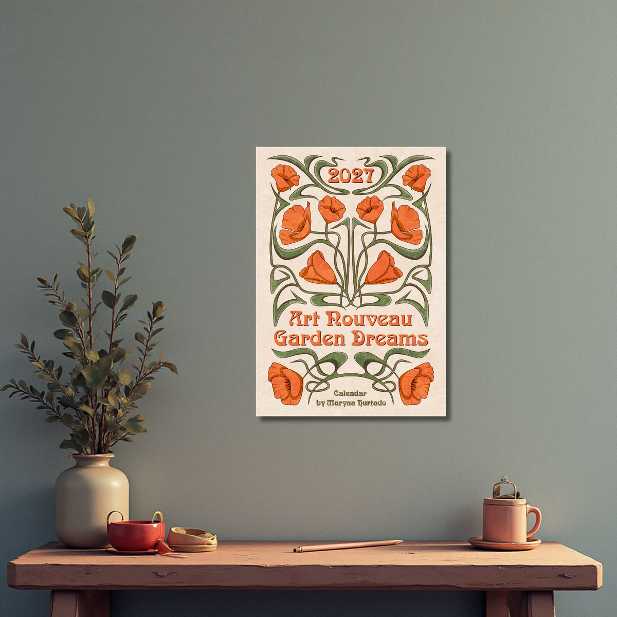 Art Nouveau Garden Dreams calendar hanging on a gray-green wall above a shelf with a plant