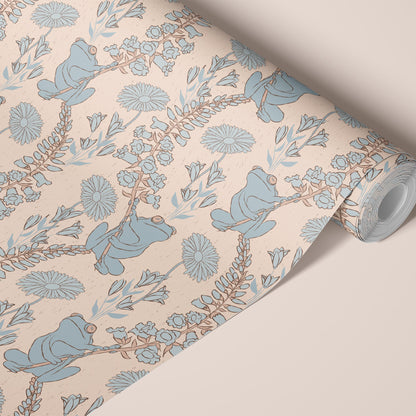 A roll of wallpaper with a whimsical pattern of blue frogs, floral elements, and delicate branches on a warm cream background, shown on a neutral beige surface.