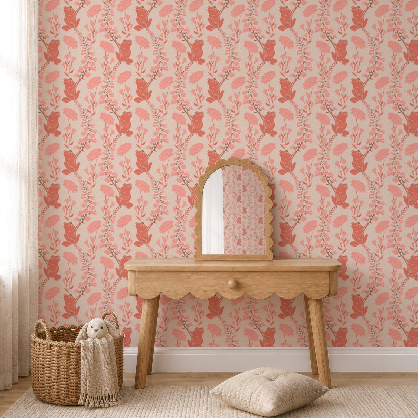 A styled interior scene showing a wall covered in pink and coral frogs wallpaper, with a light wood console table, small arched mirror, woven basket, throw blanket, and a cushion on a cream rug