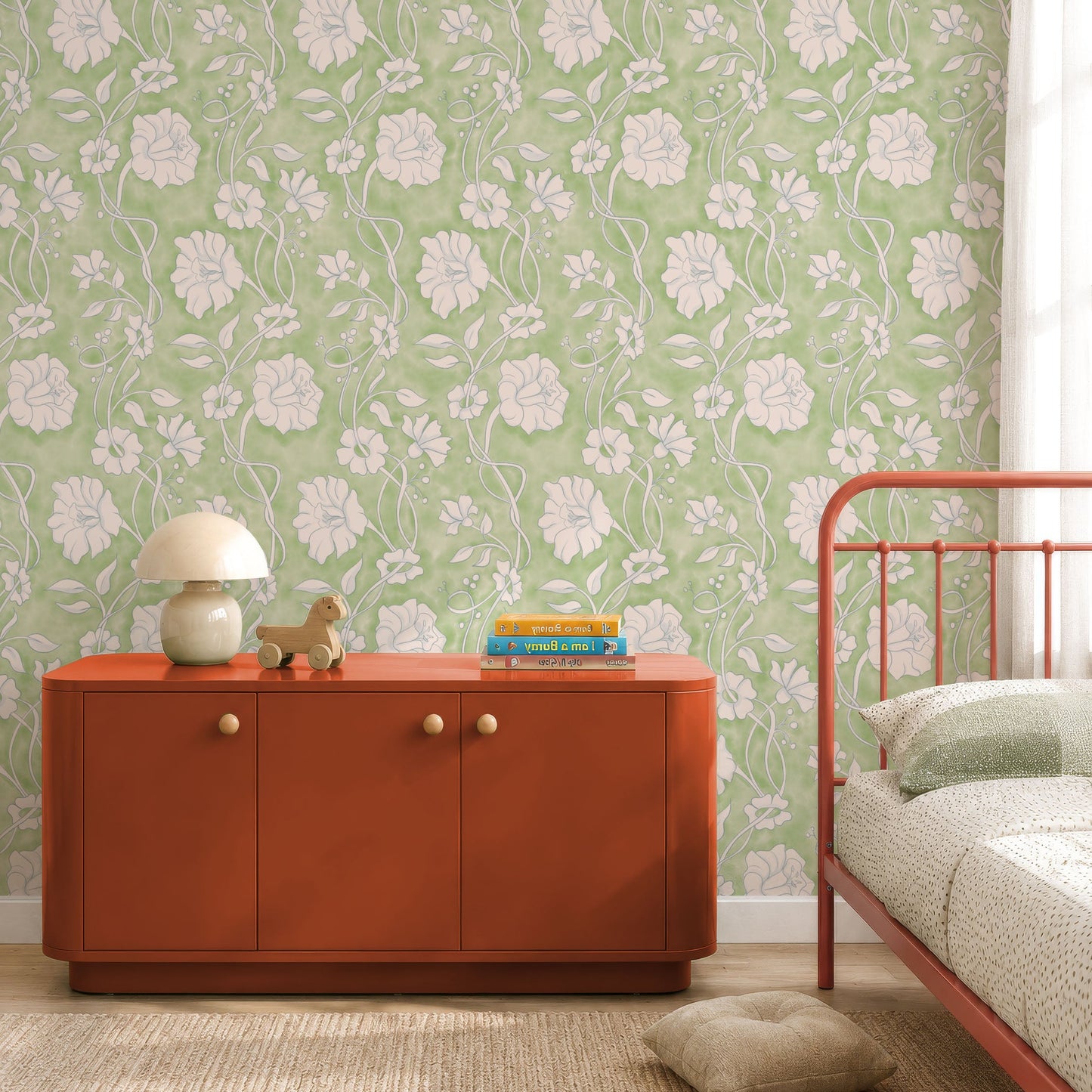 A styled bedroom corner featuring light green floral wallpaper, a rust-orange wooden sideboard with small knobs, a white table lamp, books, and a red metal bed frame.