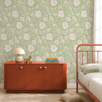 A styled bedroom corner featuring light green floral wallpaper, a rust-orange wooden sideboard with small knobs, a white table lamp, books, and a red metal bed frame.