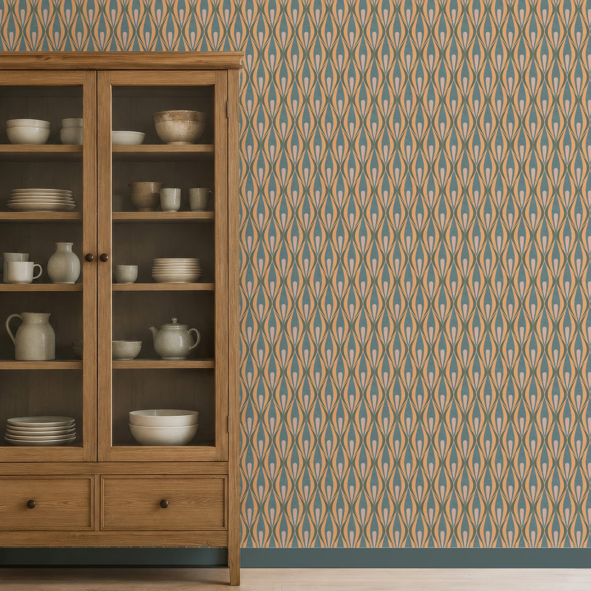 A wooden display cabinet stands against a wall covered in 70s style wallpaper with beige, blue, and brown ogee motifs. The cabinet has glass upper doors and two lower drawers, with white dishes and ceramic vessels on the shelves.