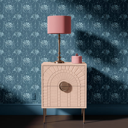A pink nightstand with a textured geometric front and gold legs, styled with a pink lamp and small pink container against a blue patterned wallpaper background.