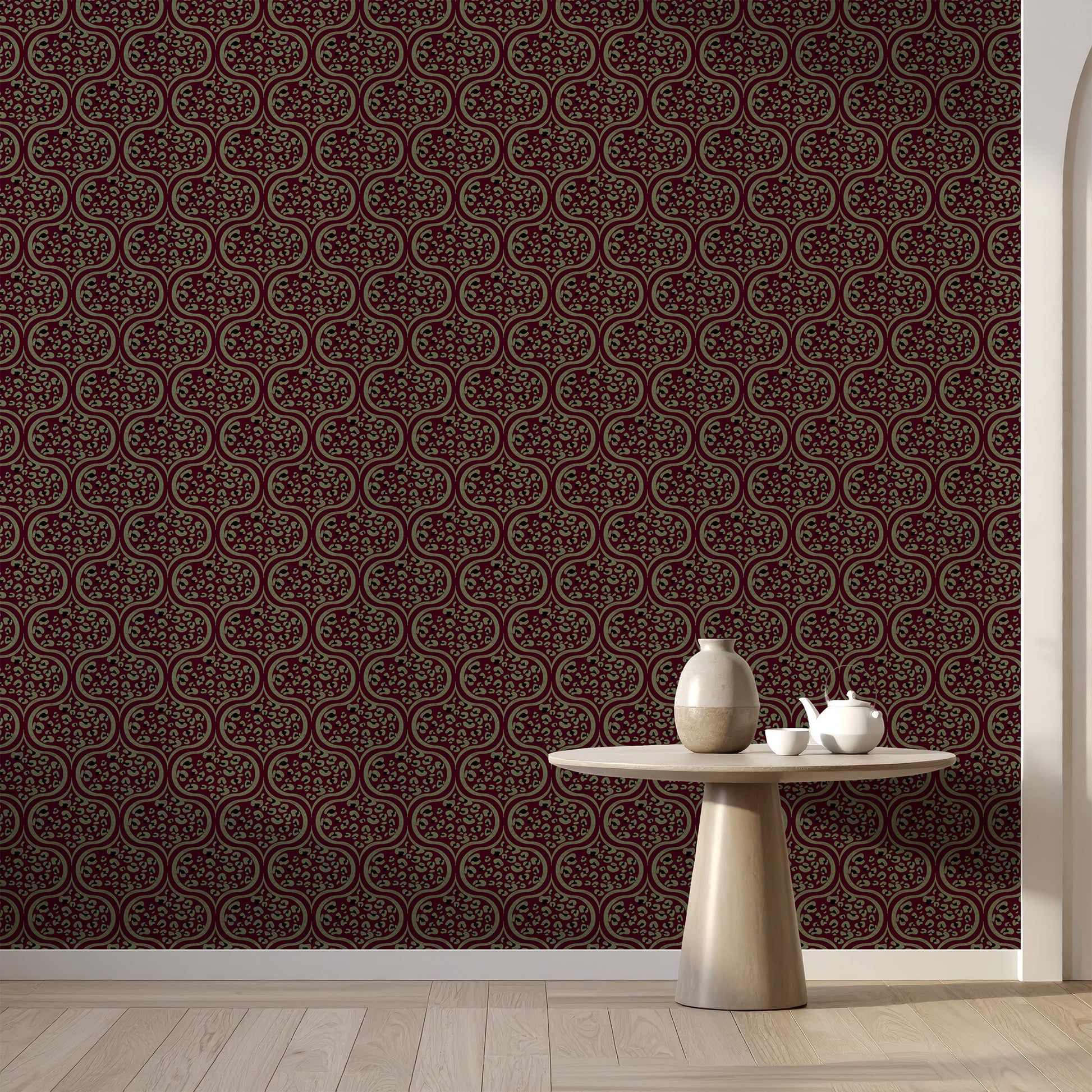 Dark brown and sage ogee patterned wallpaper shown on a wall in a styled room setting with a light wood floor and a small round table holding a vase and teapot.