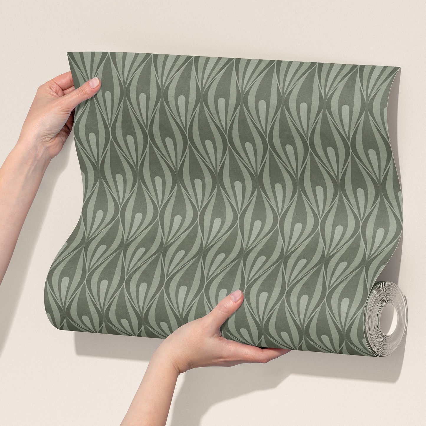 A person's hands hold up a roll of wallpaper with a repeating vertical ogee pattern in muted sage green and gray against a plain beige wall.