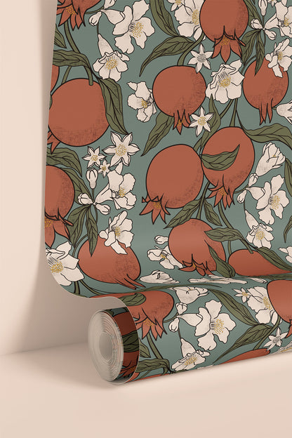 A roll of wallpaper with a repeating pattern of pomegranates, white blossoms, and green leaves on a muted sage green background, shown standing against a light beige wall.